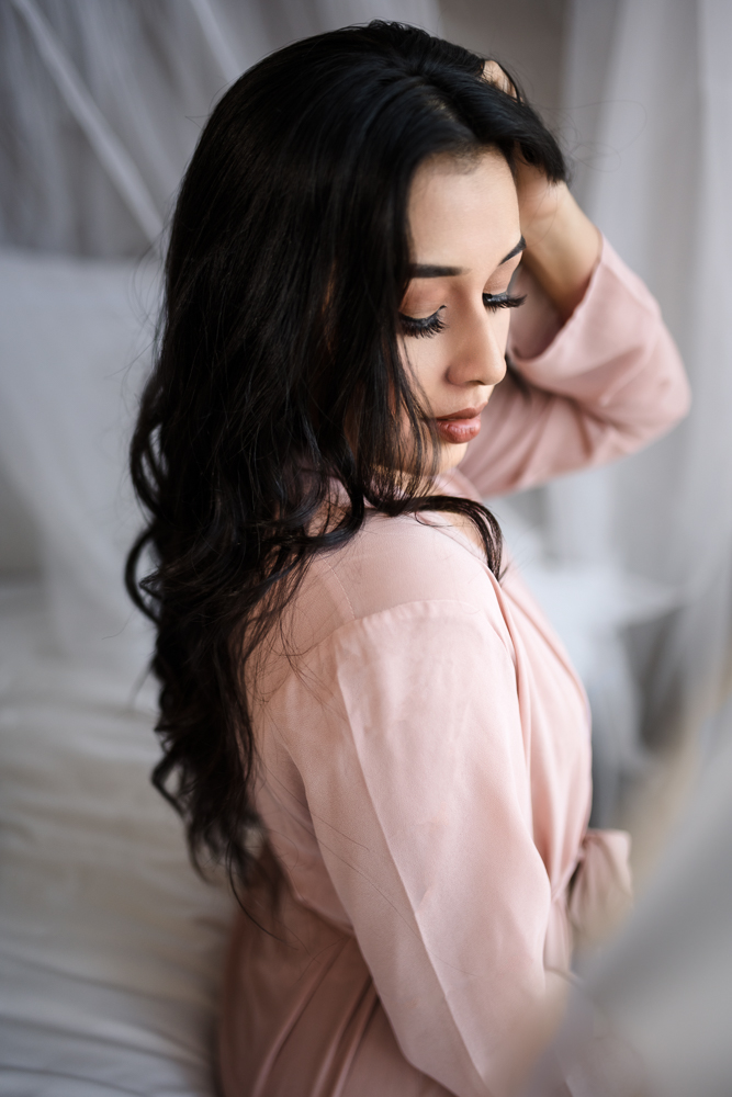 A woman in a pink robe looking down and brushing her brunette hair. There is white sheets in the background