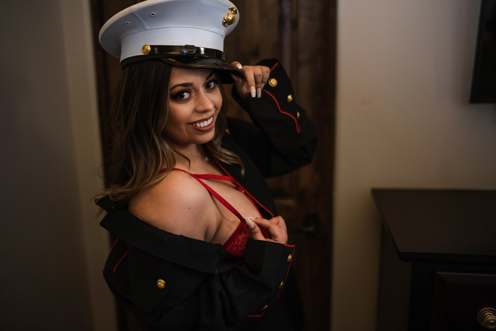 A beautiful woman tips her hat and looks playfully at the camera while wearing a red teddy and a military jacket as her boudoir outfit.