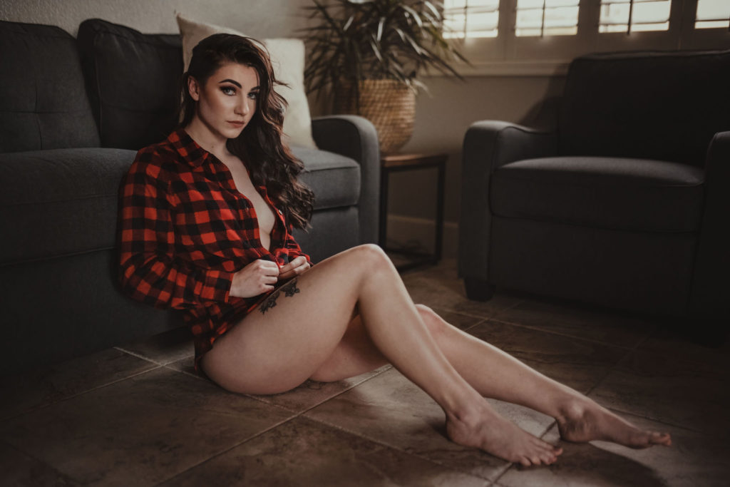 A beautiful woman who is only wear a red checkered button up flannel as her boudoir outfit. Her blue eyes are looking at the camera. The sunlight is peering though lighting her hair and make up. The editing style is moody.