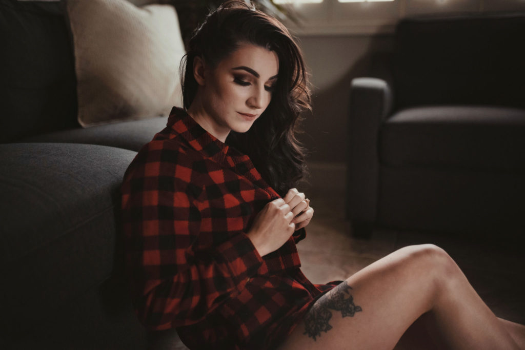 A young woman with a tattoo on her leg is wearing panties and is looking down buttoning up a red checkered flannel. 