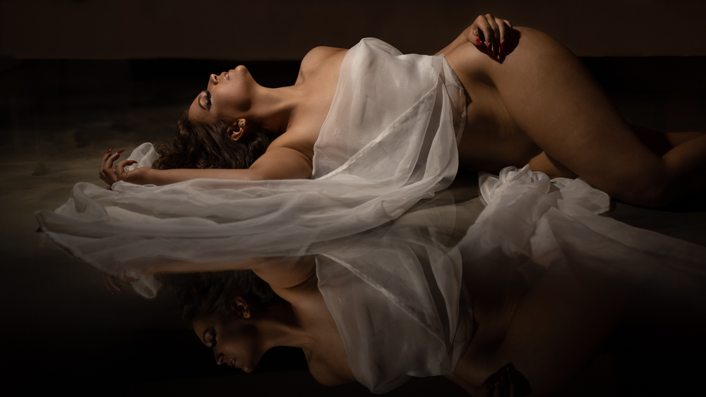 A gorgeous woman poses wrapped in a white tulle sheet like a goddess. The floor is a reflective marble that creates a mirror image of her. The woman's eyes are closed and she cranes her neck upwards as if basking in the spotlight.