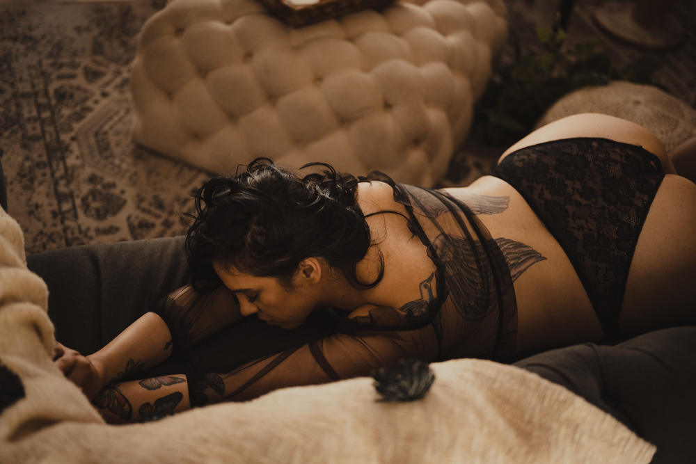 A woman in sexy black lingerie is scratching over a couch with golden light on her. The tattoos are mesmerizing and she looks confident as she does it