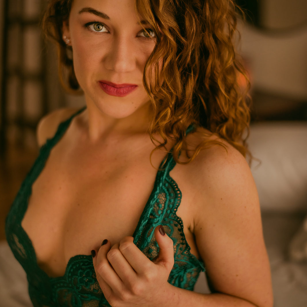 A woman is wearing green lingerie and running her fingers through the straps gazing at the camera while she sits on a bed in her bedroom.